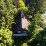 Das Bild zeigt eine Draufsicht auf ein Haus mit dunklem Dach, von Bäumen umgeben, in sonniger Umgebung. Es gibt keine deutlichen Zeichen, dass das Haus ein Abrisshaus ist, es erscheint in gutem Zustand.