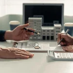 Zwei Personen sitzen an einem Tisch und diskutieren über ein Bauprojekt. Sie verwenden architektonische Modelle, Pläne, einen Taschenrechner und einen Laptop, um Details zu überprüfen.
