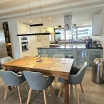Ein moderner Essbereich in einem offenen Wohnkonzept mit angeschlossener Küche. Der Tisch aus Holz mit Platzsets und sechs Stühlen zentriert den Raum. Links eine Edelstahl-Kühlschrank. Rechts eine Küchenzeile mit Hängeschränken und Küchengeräten.