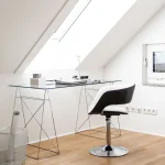Ein moderner Arbeitsplatz unter einer Dachschräge mit einem Glas-Schreibtisch auf filigranem Metallgestell, einem schwarzen Designerstuhl, einem Laptop, einer Lampe und einer transparenten Papierkorb. Parkettboden und ein Dachfenster sind ebenfalls zu sehen.