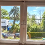 Blick aus einem weißen Fenster auf ein modernes Haus, Bäume und einen sonnigen See.
