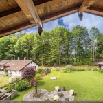 Vom Balkon aus sieht man einen idyllischen Garten mit gepflegtem Rasen, umgeben von Bäumen. Drei Schafe grasen im Vordergrund. Auffällig sind ein rotes Dachhaus und eine kleine Hütte. Der Blick wird durch Holzbalken gerahmt.