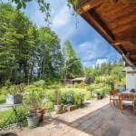 Das Bild zeigt eine idyllische Terrasse eines Landhauses am Tegernsee mit Holzmöbeln und einem Grill. Der gut gepflegte Garten ist umgeben von hohen Bäumen. Helle, natürliche Farben dominieren die Szenerie.