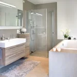 Modernes Badezimmer: Holzwaschtisch mit Aufsatzbecken, große Spiegel, Glasduschabtrennung, eingelassene Badewanne, graue Fliesen. Deko: Kerzen, Pflanzen, Handtücher. Leuchten im Spiegel integriert.