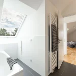 Das Bild zeigt ein modernes Bad en suite mit direktem Zugang zu einem Schlafzimmer. Im Bad sind eine Badewanne mit einem Handtuch, weiße Wände und graue Bodenfliesen sichtbar. Über der Badewanne befindet sich ein Dachfenster, das den Blick in den Himmel freigibt.