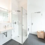 Modernes Badezimmer mit großer, gläserner Duschabtrennung, weißem Waschbecken, Spiegel mit Beleuchtung, grauen Bodenfliesen und einer Waschmaschine. Ein Korb und ein Heizkörper sind ebenfalls zu sehen. Keine Badewanne vorhanden.