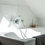 Ein helles Badezimmer mit einer weißen Badewanne unter einer Dachschräge. Die Armatur ist modern, aus Chrom. Auf dem Rand stehen Pflegeprodukte und ein Naturschwamm, was auf ein entspannendes Badevergnügen hindeutet.