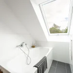 Modernes Badezimmer mit weißer Badewanne unter einem Dachfenster, durch das man den blauen Himmel und Bäume sieht. Weiße Fliesen an den Wänden, dunkelgraue Fliesen am Boden und ein Korb mit Handtüchern neben der Wanne.