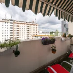 Blick von einem gemütlichen Balkon mit Markise und roten Stühlen auf ein großes Mehrfamilienhaus und eine Baustelle