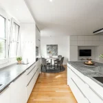 Das Bild zeigt eine moderne, helle Küche mit weißen Schränken und dunkler Arbeitsplatte. Links im Bild ist ein Fenster. Durch die offene Raumgestaltung sind ein Essbereich mit rundem Teppich und grauen Stühlen sowie ein Wohnzimmer mit Fernseher erkennbar.