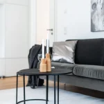Ein Wohnzimmer mit modernem Interieur: rechts graues Sofa mit Kissen, links kleiner runder Tisch mit Kerzenhaltern und einer Glühbirne. Hintergrund zeigt weiße Regale, ein Gemälde und einen Fernseher.