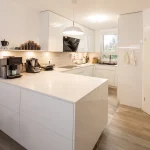 Moderne weiße Einbauküche mit Kochinsel und hochwertigen Küchengeräten in einem hellen Wohnraum.