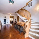 Das Bild zeigt den Innenraum eines Landhauses mit einer eleganten Holztreppe, die einen geschwungenen Verlauf nimmt. Es gibt Elemente aus Holz und eine rustikale Einrichtung, inklusive einer hölzernen Anrichte und Hirschgeweih-Dekoration an der Wand.