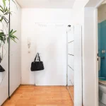Heller Flur einer Wohnung mit weißer Eingangstür, Holzboden, Schuhschrank und Blick in ein blau gefliestes Badezimmer