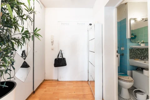 Heller Flur einer Wohnung mit weißer Eingangstür, Holzboden, Schuhschrank und Blick in ein blau gefliestes Badezimmer
