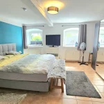 Ein helles Gästezimmer mit einem großen Bett, blau-türkiser Wand, zwei Fernsehern, einer Standventilator und Spiegel. Es gibt auch zwei Fenster, Teppiche und eine Deckenlampe. Der Raum wirkt modern und einladend.