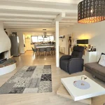Das Bild zeigt den Wohnbereich einer Wohnung mit offenem Grundriss. Im Vordergrund ist ein großer Kamin. Der Raum ist modern eingerichtet mit einem grauen Sofa, Sesseln und einem weißen Couchtisch. Im Hintergrund ist die Küche.