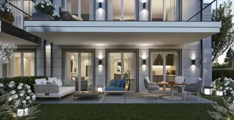 Das Bild zeigt eine moderne Gartenansicht eines zweistöckigen Hauses bei Abenddämmerung. Im Vordergrund ist ein gepflegter Rasen mit Blumenbeeten und Gartenmöbeln zu sehen. Spotlights beleuchten die Fassade stilvoll.