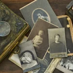 Geschichte, Historie und Persönlichkeiten: Auf dem Bild sind alte Fotografien und Gegenstände zu sehen, die an historische Zeiten erinnern. Dazu gehört ein abgenutztes Buch, eine Lupe, eine Holzbox und vertrocknete Rosen. Die Fotos zeigen Porträts verschiedener Personen aus einer vergangenen Epoche.