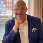Harald König, Immobilienexperte bei GoldHaus Immobilien, lächelt freundlich vor einem Fenster mit Stadtblick.