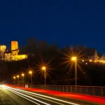 Auf dem Bild ist eine nächtliche Szene mit einer beleuchteten, mittelalterlichen Burg und einer Kirche auf einem Hügel. Im Vordergrund sieht man eine gestrahlte Straße mit Lichtspuren von Autos und mehreren Straßenlaternen. Der klare Sternenhimmel erstreckt sich darüber.