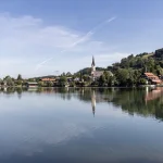 Das Bild zeigt den Schliersee in Deutschland mit ruhigem, spiegelglattem Wasser und einer Reflexion des Himmels sowie der umgebenden Landschaft. Zu sehen ist eine Kirche mit markantem Turm, dahinter hügelige Landschaft und bewaldete Hänge. Wohnhäuser verteilen sich am Ufer.