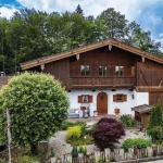 Ein typisches, großes alpenländisches Holzhaus in Tegernsee mit verschindelter Fassade, Balkon, weißen Fensterrahmen und einer großen Garage. Vor dem Haus befindet sich ein gepflegter Garten mit Ziersträuchern und einem Kiesweg.