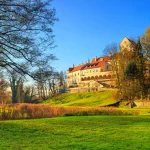 Das Bild zeigt ein großes, historisches Haus auf einem Hügel, umgeben von einer üppigen Parklandschaft. Das Haus besitzt eine Mischung aus gotischen und Renaissance-Elementen, mit einem roten Ziegeldach und weißen Fassaden. Vor dem Haus erstreckt sich eine große Wiese, durchzogen von einem Pfad und herbstlich gefärbter Vegetation. Dies könnte eine ideale Immobilie für jemanden sein, der in Seefeld ein Haus verkaufen oder kaufen möchte.