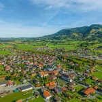 Panoramablick auf Unterammergau, ein idyllisches Dorf umgeben von bewaldeten Bergen. Es zeigt dicht bebaute Bereiche mit traditioneller Architektur, durchzogen von Straßen und umgeben von Feldern und Wiesen. Ideal für Immobilienmakler.