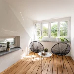 Das Bild zeigt eine Innenloggia mit schrägen Wänden und Fenstern mit Blick auf grüne Bäume. Ein Holzboden und zwei moderne schwarze Drahtstühle um einen weißen Rundtisch sind zentral platziert. An der Wand ist ein Einbau-Kamin.