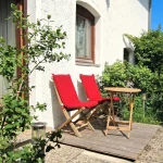 Zwei Holzklappstühle mit roten Auflagen und ein kleiner Holztisch stehen auf einer kleinen Holzterrasse vor einer weißen Hausfassade.