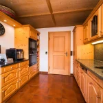 Rustikale Küche in einem Haus am Tegernsee mit holzvertäfelten Wänden und Decke, Holzschränken, dunklem Granitarbeitsplatten, modernen Küchengeräten und einer runden Wanduhr.