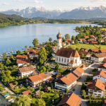 Drohnenaufnahme des Seedorfes Hechendorf mit Pfarrkirche, Pilsensee-Ufer und Alpenpanorama.