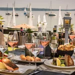 Auf dem Bild ist eine elegante Tischdekoration in einem Restaurant am Ufer zu sehen. Auf dem Tisch stehen diverse Gerichte wie gebratener Fisch, Salate und Brotkorb, ergänzt durch mehrere Gläser mit Roséwein. Im Hintergrund sind zahlreiche Segelboote auf einem See zu erkennen.