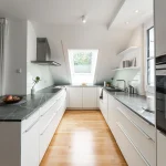 Moderne Einbauküche unter Dachschrägen mit weißen Schränken, dunkler Arbeitsplatte aus Naturstein, Holzparkettboden, Einbaugeräten von Siemens und zwei Fenstern, die für natürliches Licht sorgen.