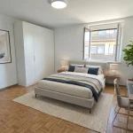 Ein modernes Schlafzimmer unter einem schrägen Dach mit Parkettboden. Es hat ein großes Bett mit grauer Bettwäsche, Nachttisch mit Lampe, eine Sitzecke, eine offene Tür und Kleiderständer. Das Raumgefühl wird durch ein Dachfenster erhellt.