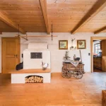 Das Bild zeigt einen Raum im traditionellen bayrischen Stil, vermutlich eine "Stubn" am Tegernsee. Ein Kachelofen steht im Zentrum, daneben ein Holztisch mit Bänken. Die Zimmerdecke besteht aus Holzbalken, und an den Wänden hängen gerahmte Bilder.