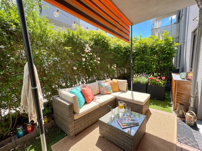 Gestaltungsbeispiel einer gemütlichen Terrasse mit Rattan-Lounge-Möbeln, Markise, Lichterkette und grüner Bepflanzung.