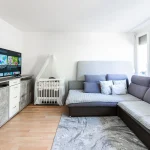 Modernes Wohnzimmer einer Wohnung mit grauer Eckcouch, Fernseher, TV-Lowboard und Babybett im Paket 2