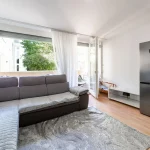 Heller Wohnbereich in Wohnung 13 mit großer grauer Couch, Holzboden und Zugang zum Balkon