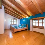 Das Bild zeigt ein rustikales Schlafzimmer in einem Holzhaus am Tegernsee. Charakteristisch sind die Holzbalken an der Decke und die Holzdielen auf dem Boden. Die Wände sind in einem kräftigen Blau gestrichen. Das Doppelbett ist mit einer blau-weißen Bettwäsche bezogen, und es gibt eine Stehlampe neben dem Fenster, das mit weißen Vorhängen versehen ist.